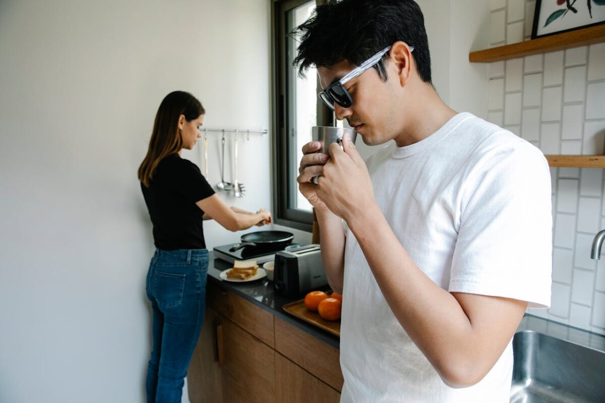 Offres A man wearing sunglasses drinks while a woman cooks in a modern kitchen setting.